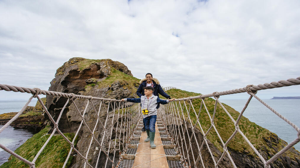 Due persone camminano su un ponte di corda sopra l'acqua vicino a una scogliera e una collina verde.