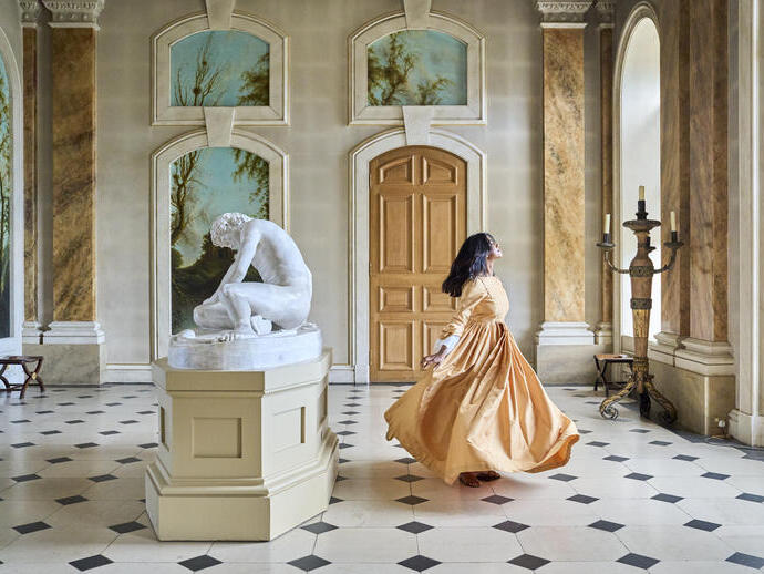 Femme en longue robe tournant dans une grande salle avec une statue en marbre, des colonnes et des murs décorés.