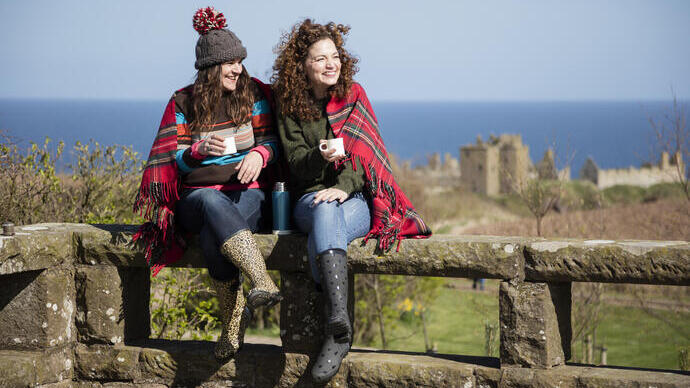 Dos mujeres con bebidas calientes, sentadas en un muro de piedra cerca de un castillo