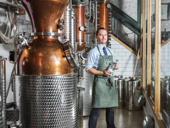 Uomo con grembiule verde in piedi accanto a un alambicco di rame in una distilleria di gin