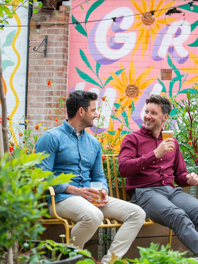 Two men sat on a bench in a garden with a drink