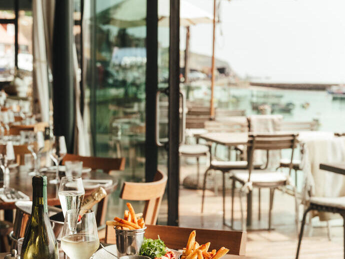 Seaside restaurant with tables set for dining, plates of lobster, fries, salad, wine glasses, and ocean view through glass walls.
