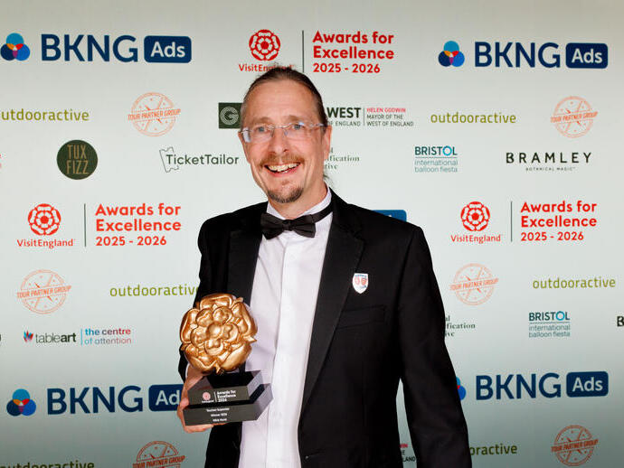 Person in formal attire holding a trophy at an awards event with branded backdrop for Awards for Excellence 2025-2026.
