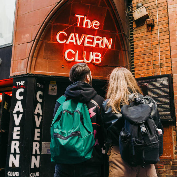 Two people with backpacks stand outside The Cavern Club, a famous brick music venue with neon signage in Liverpool.
