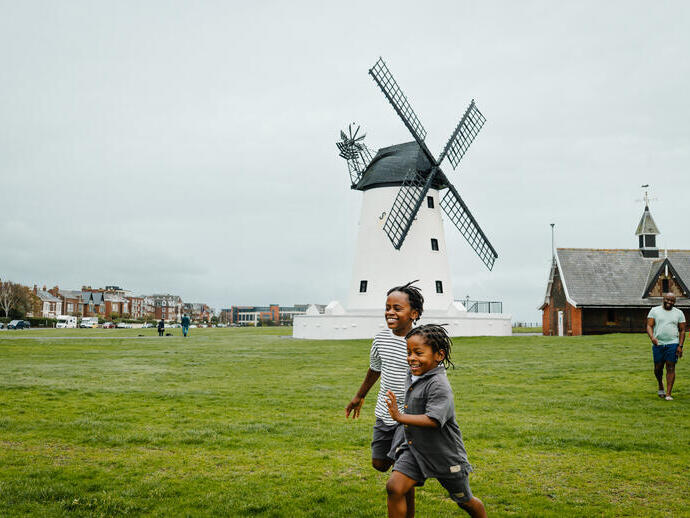 Children run on a green field near a traditional white windmill and buildings, with adults walking in the background on a cloudy day.