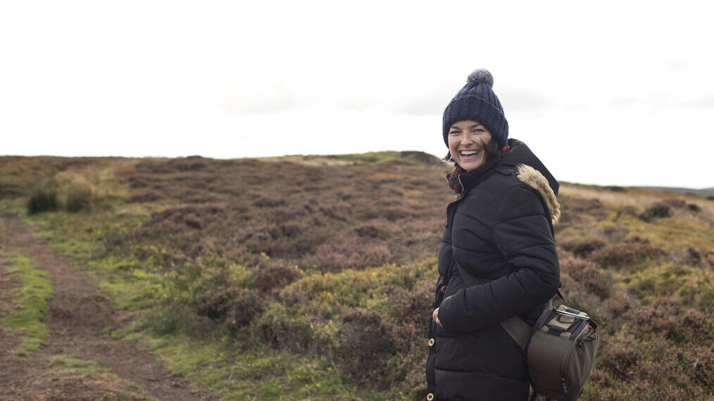 Junge Frau in einem dicken Mantel und Hut, die auf einem Weg in einem Moor entlanggeht