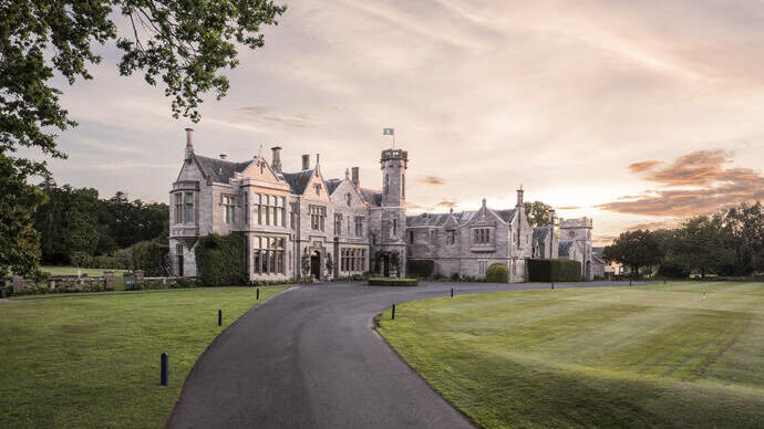 Large historic manor house with stone facade, surrounded by green lawns and trees, under a dramatic sunset sky.