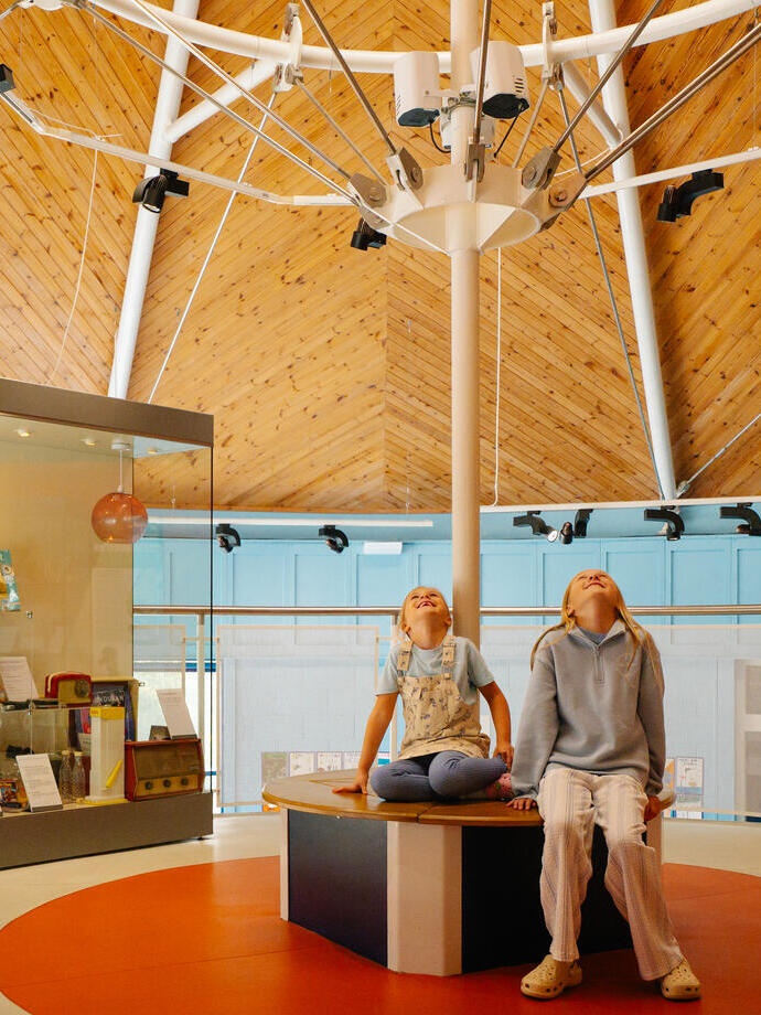 Two young girls sitting in the centre of a room looking up to the ceiling in a museum