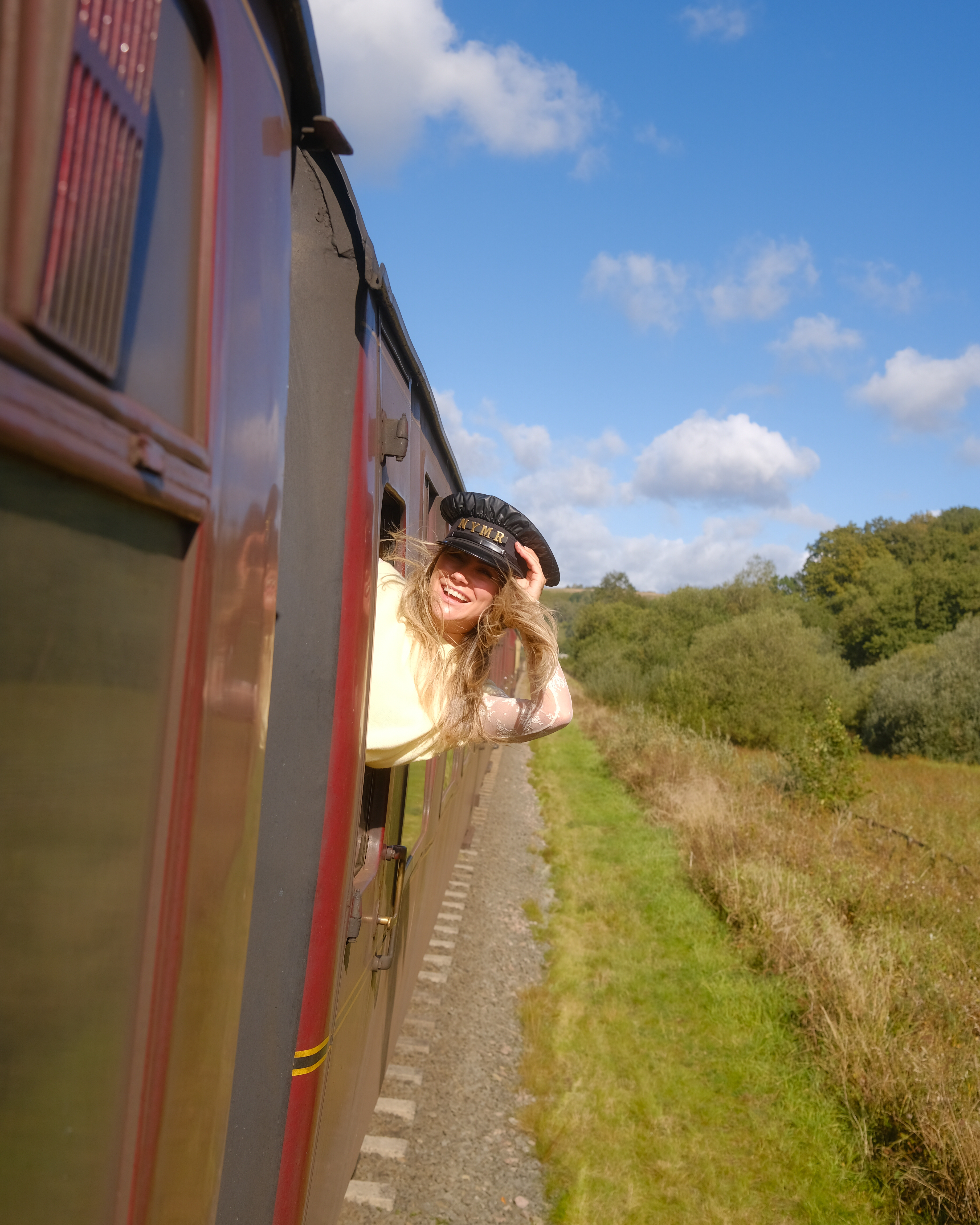Person leaning out of a traditional train carriage on a sunny day, smiling, with rural landscape and blue sky in the background.