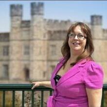 Person in a bright pink outfit standing in front of a historic stone castle with towers, on a sunny day.