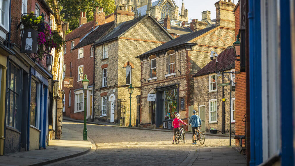 Two cyclists walking uphill with bikes in Lincoln, with Lincoln Castle in the background