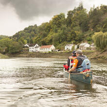 Two people canoe on a calm river with houses and forested hills under a cloudy sky in the background.