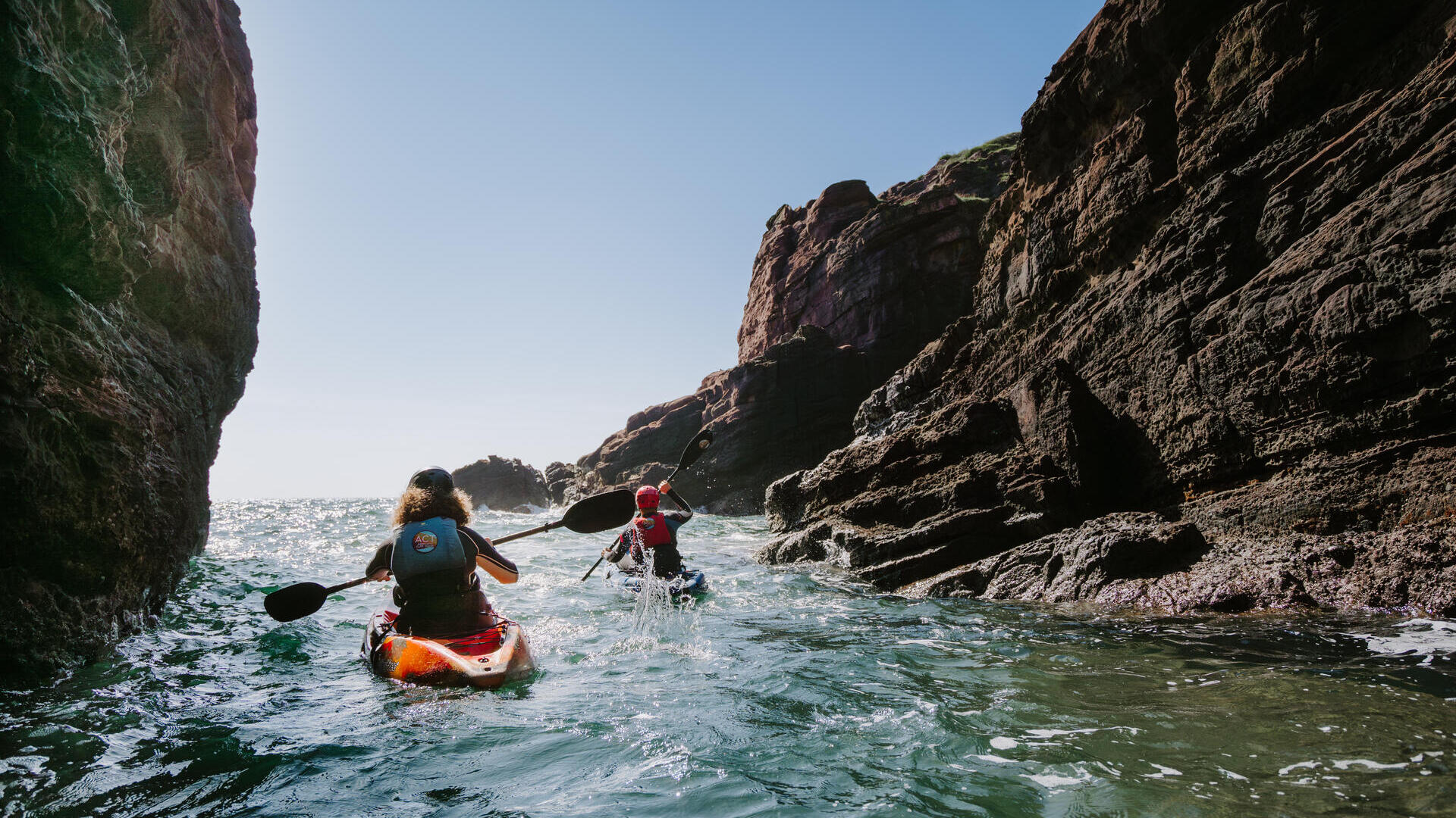 شخصان يتجدفان بقوارب الكاياك بين منحدرات صخرية على البحر تحت سماء صافية.