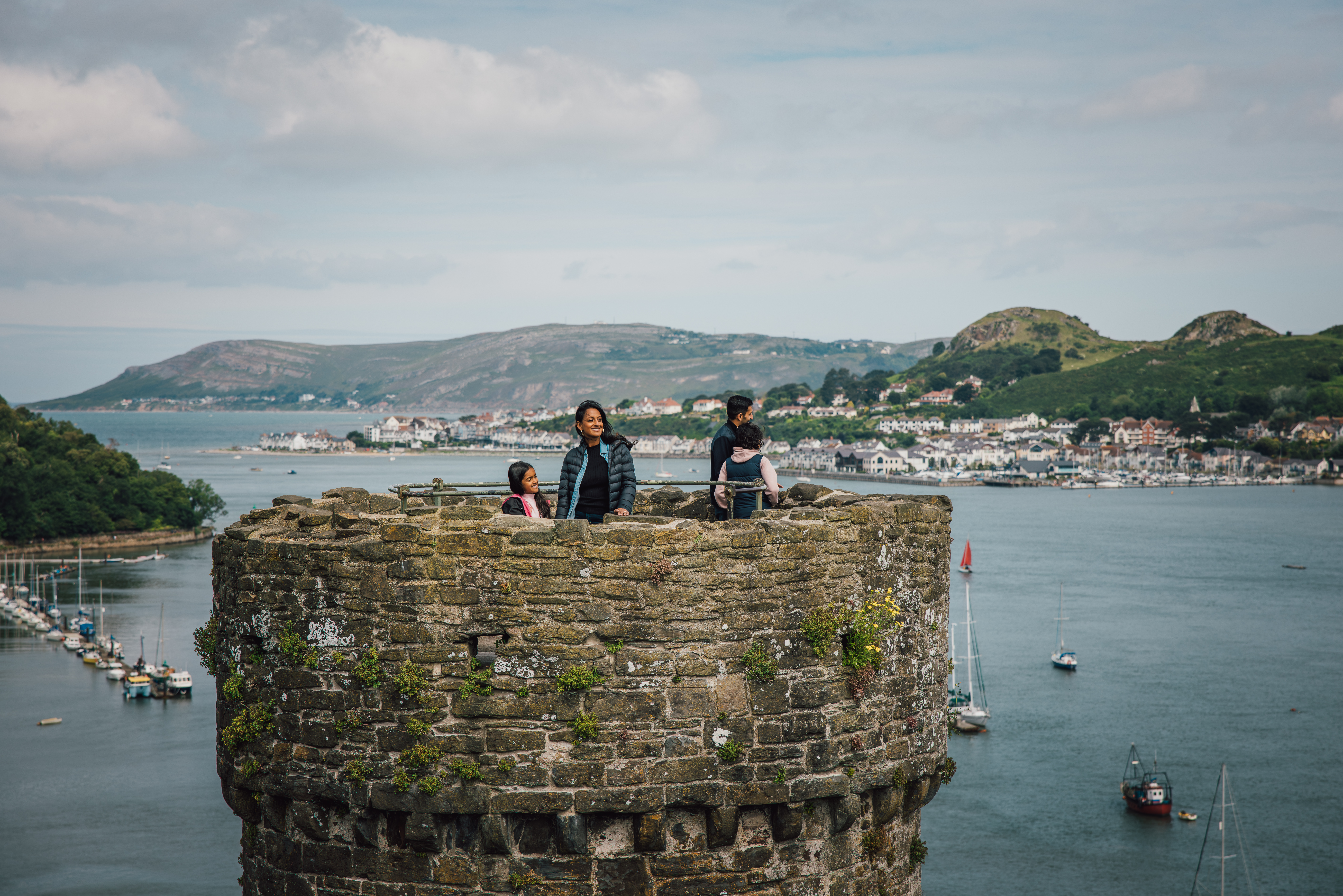People standing atop an old stone castle tower overlooking water, sailboats, and a coastal town with hills in the background under a cloudy sky.