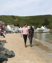 Two people walking along a sandy beach with the ocean to one side and pubs and shops on the other side.