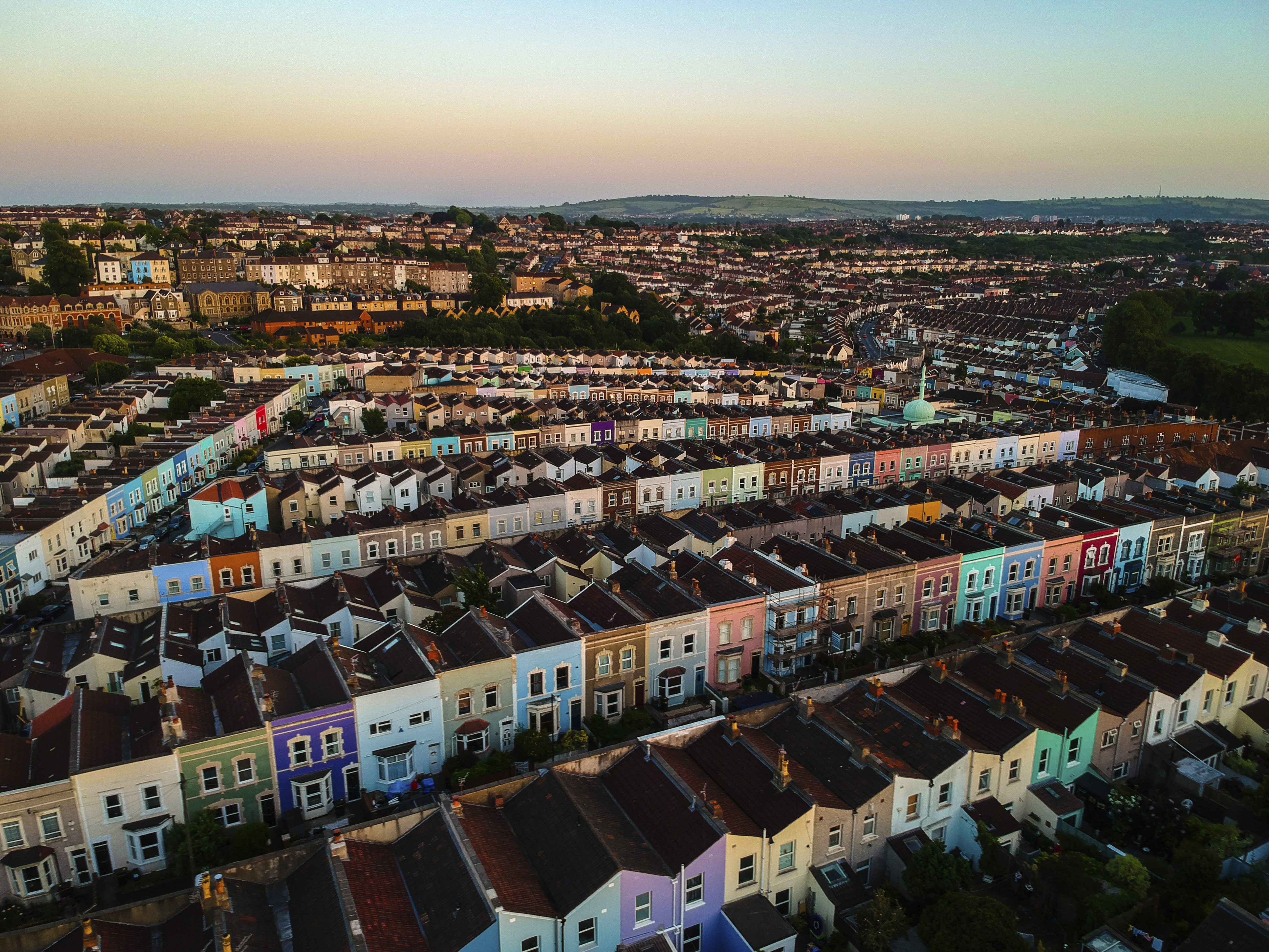 Aerial view of a residential area with rows of colourful terraced houses, greenery, and cityscape under a clear sky.