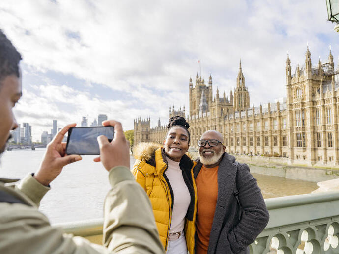 Un hombre fotografiando a una pareja madura con un gran río y edificios emblemáticos al fondo.