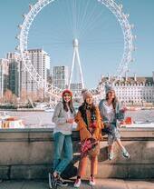 Drei Frauen sitzen auf einer Mauer vor einem großen Fluss, Gebäuden und einem Riesenrad