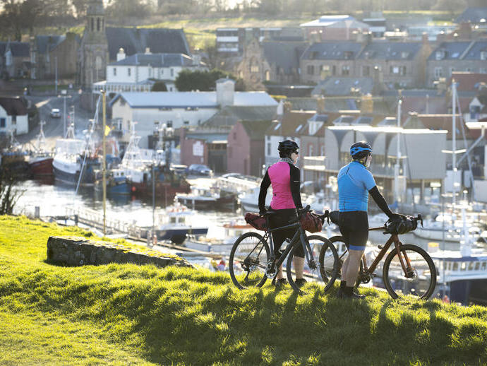 Two cyclists with gear overlook a harbor with boats and historic stone buildings under sunlight, suggesting outdoor adventure and scenic town views.