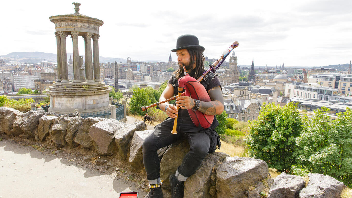 Man playing the bagpipes sat on a rock on a hill overlooking a city