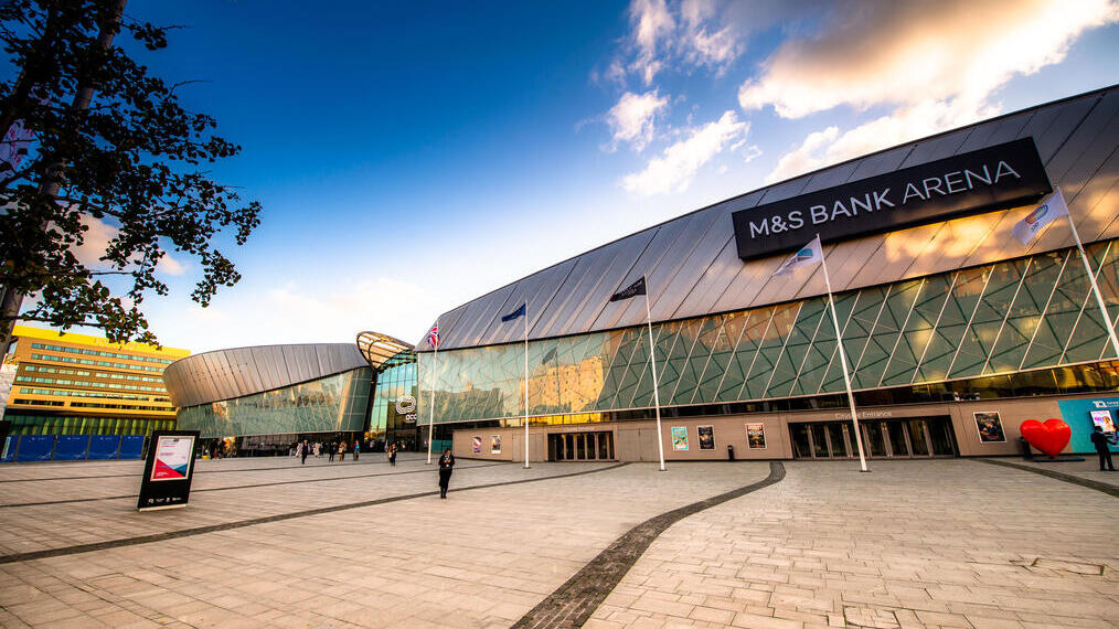 Modern glass-fronted M&S Bank Arena with flags, posters, people, and a large open plaza under a blue sky with clouds.