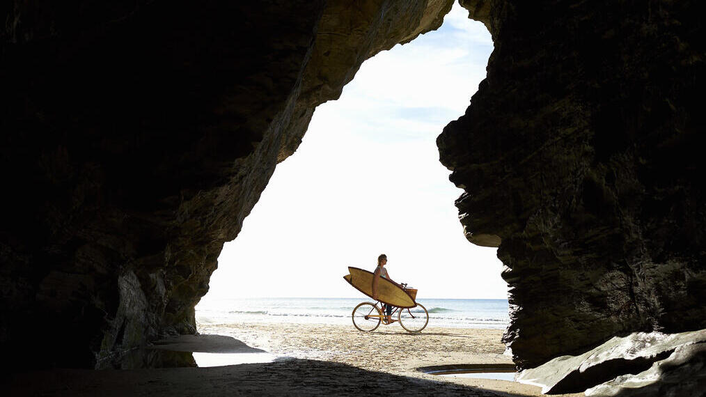 Persona en bicicleta con tabla de surf en la playa, vista desde el interior de una cueva.