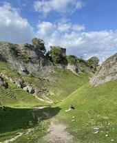 Une vallée par une belle journée d'été, avec un couple se reposant sur l'herbe. Un château est visible à travers les arbres sur le flanc rocheux de la colline.