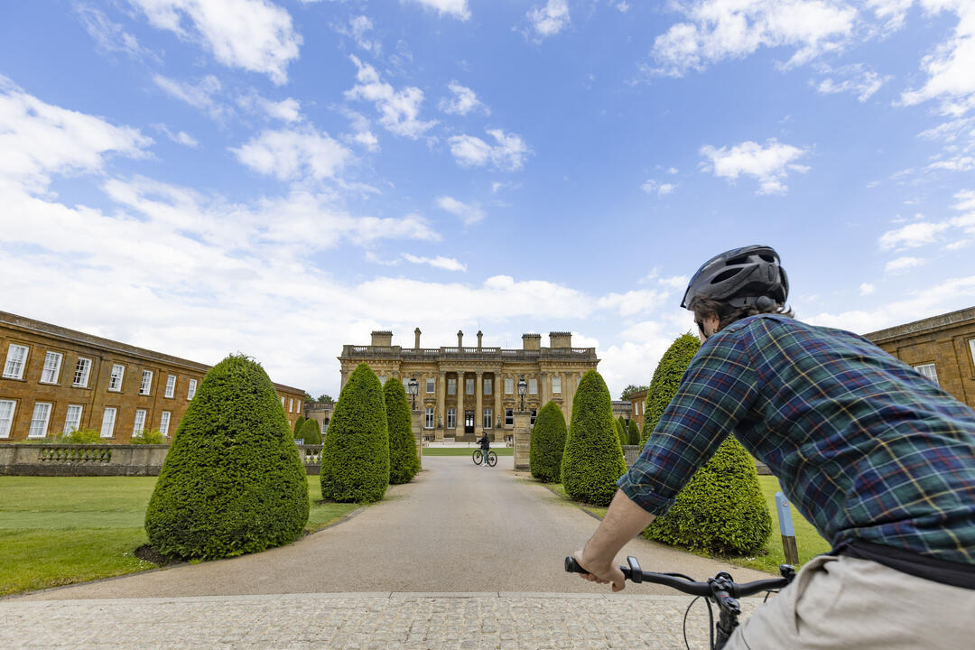 A man stands with his bike in grand gardens looking towards a stately home