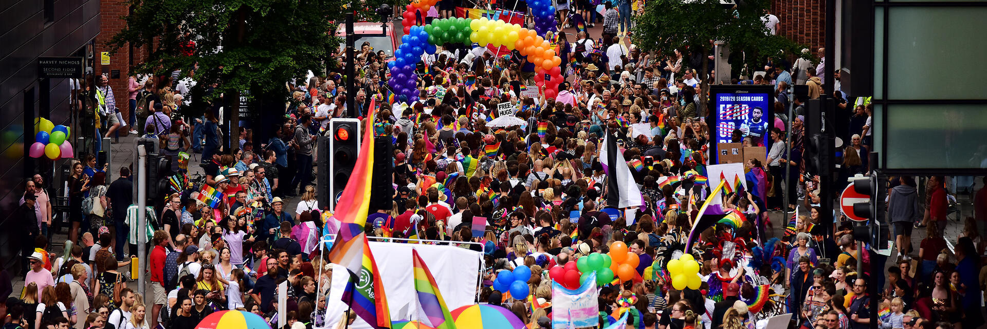 Crowd marching at a parade, holding rainbow flags, umbrellas and balloons
