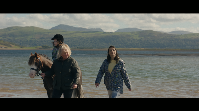 Two women walk with a child who is riding a horse on a beach with mountains beyond