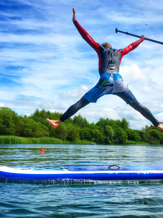 A man in a wetsuit jumps up above a paddleboard