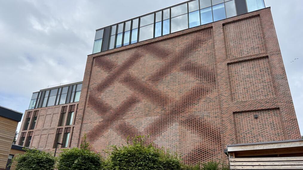 Modern brick building with geometric patterned facade, large glass windows on upper floors, and small trees in front. Sky is overcast.