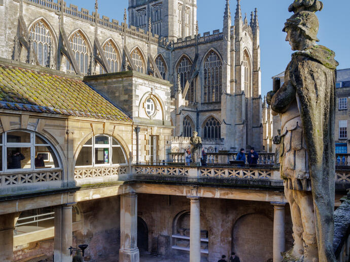 A large sculpture stands on top of roman baths ruins with an abbey beyond