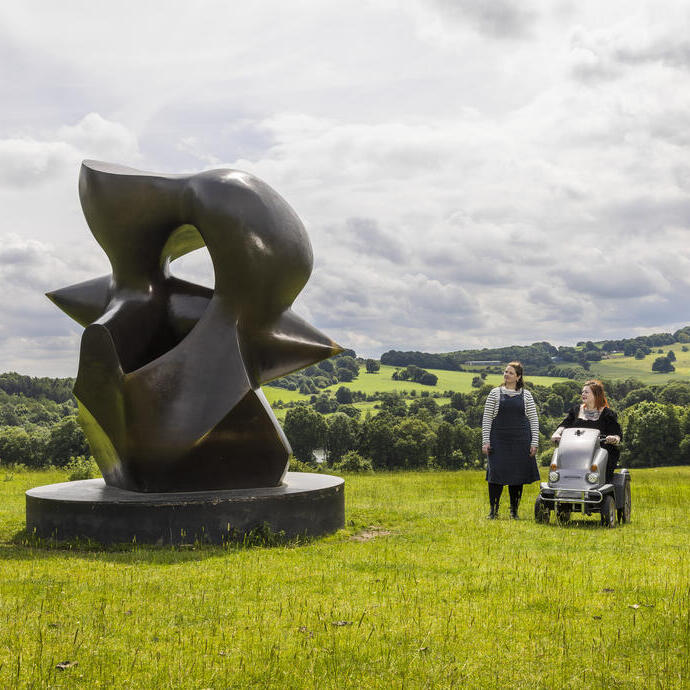 Two women look at a large sculpture set in green gardens with landscape beyond