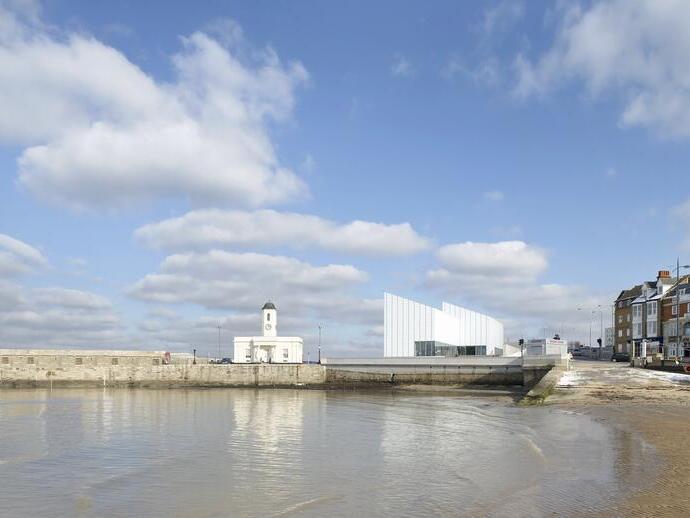 Modern building overlooking a harbour and beach, a row of townhouses in the background