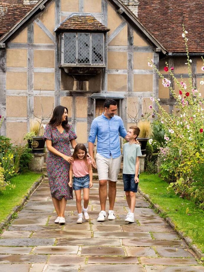 Eine Familie geht durch einen Garten vor einem historischen Haus mit schrägem Dach und blühendem Garten.