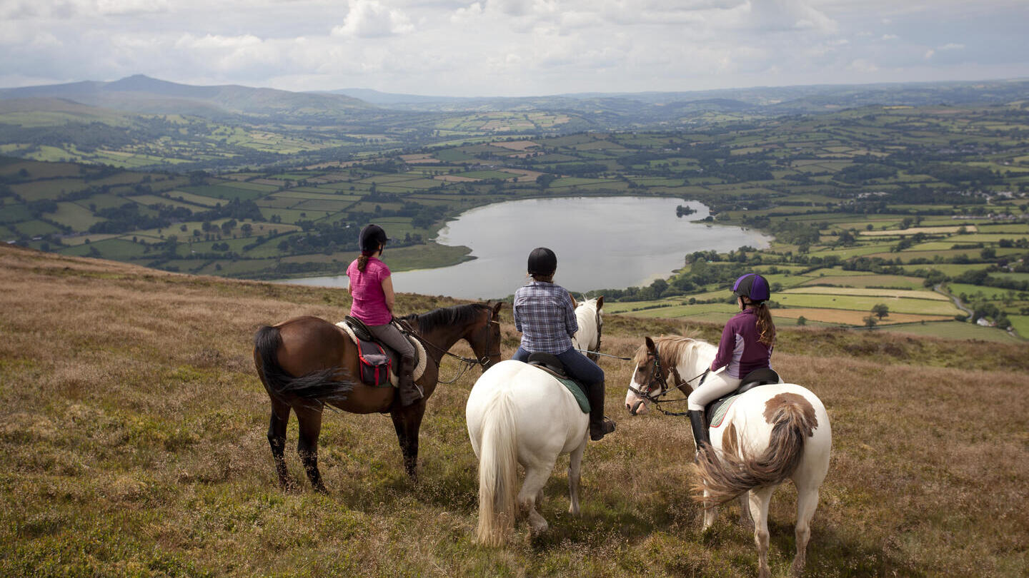 Eine Gruppe von Reitern auf einem Hügel mit Blick auf Ackerland und einen See.