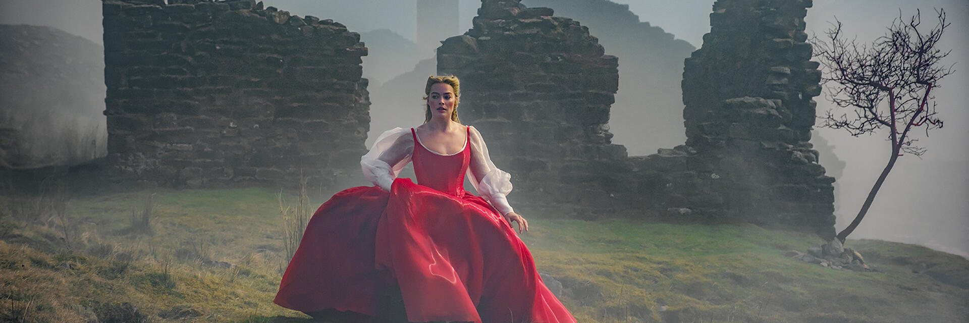 A person in a vibrant red dress stands on grass near ruins and a tree, with mist and hills in the background, evoking a dramatic scene.