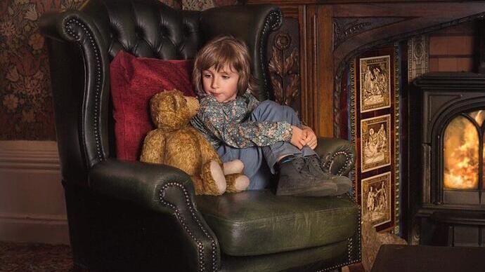 Young child sat on a green armchair in front of a fireplace with a teddy bear next to them at Criag y Nos Castle in the Brecon Beacons
