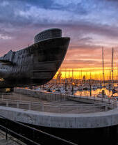 Un gran submarino junto a un puerto lleno de barcos al atardecer.