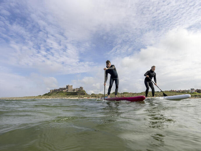 Two men on are paddleboarding in the sea with a heritage castle in the background.