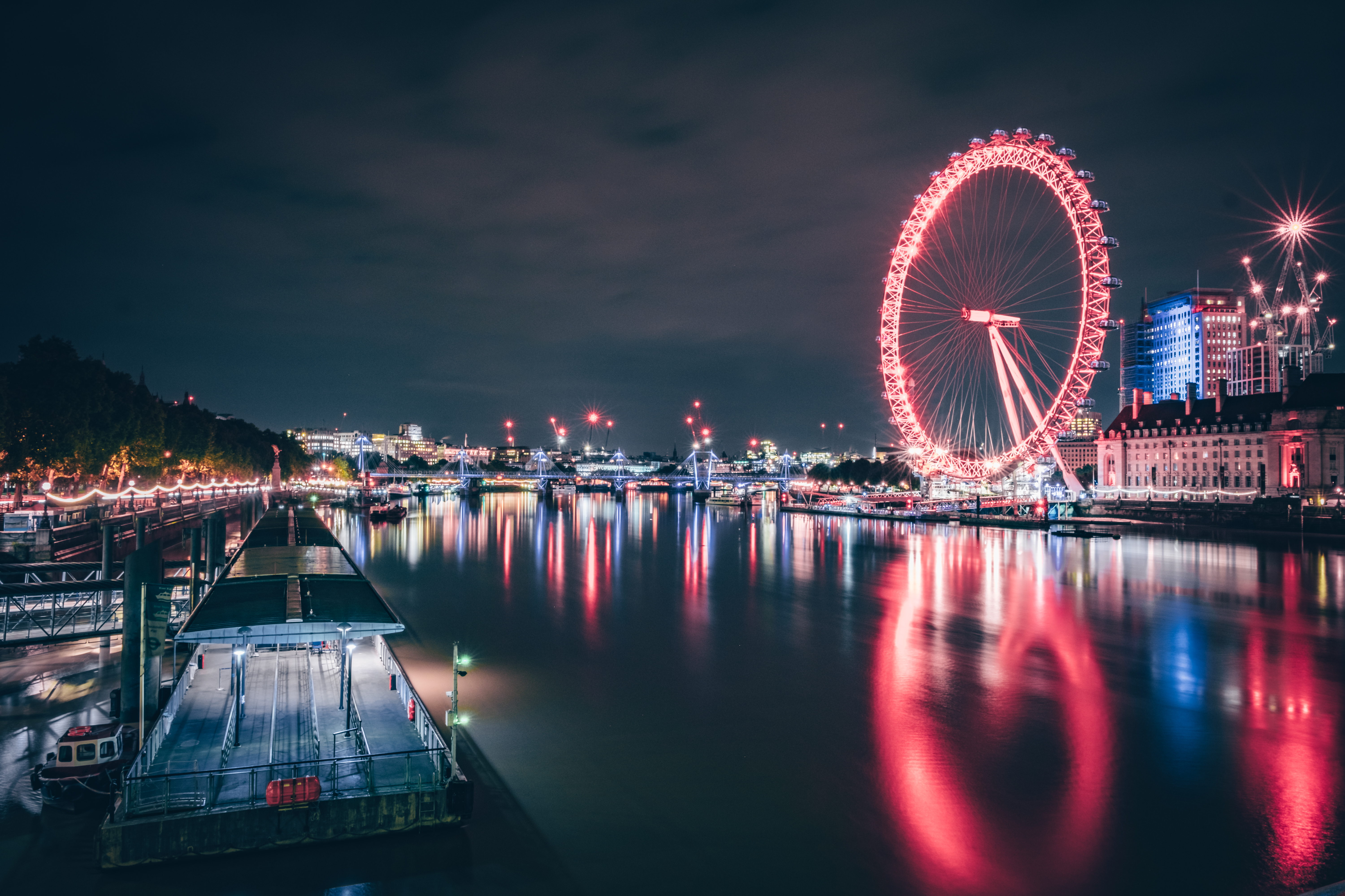 La grande roue de Londres illuminée en rouge la nuit, se reflétant sur la Tamise, avec les lumières de la ville en arrière-plan.