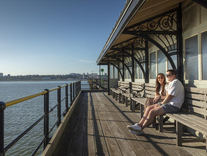 A man and a woman sit on a bench on a pier at the beach