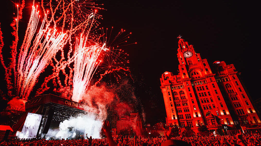 Crowds of people watching a band at a music festival as fireworks explode, in front of the Liver Building.