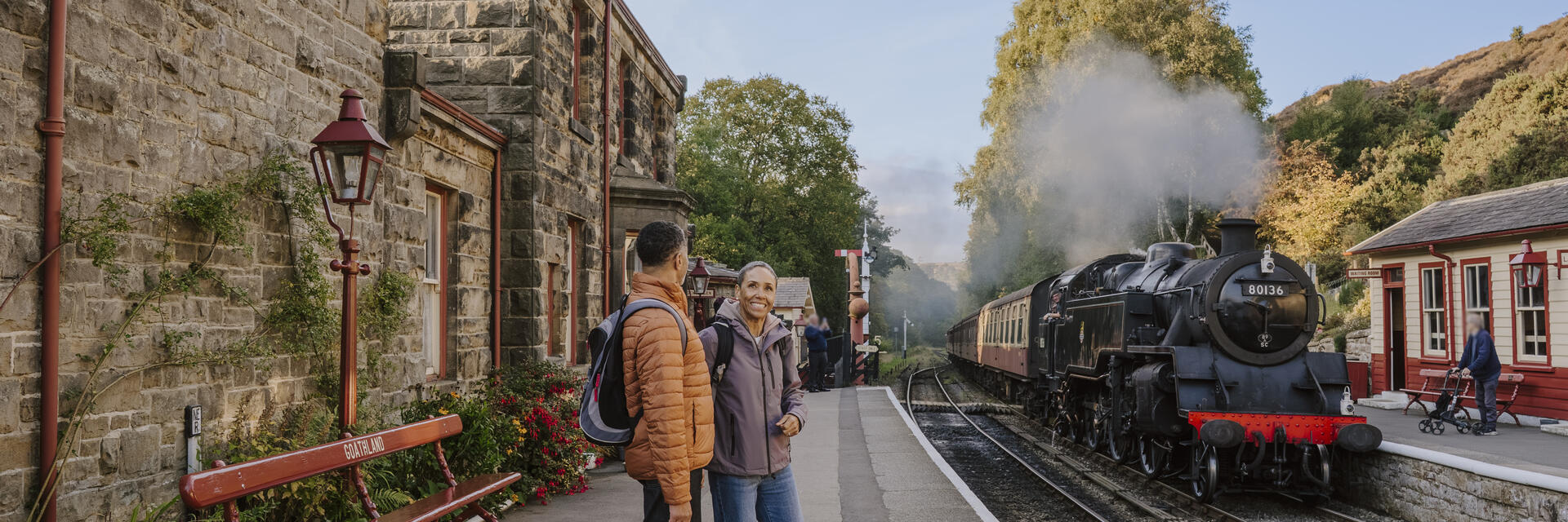 Two people stand talking on a train station platform beside an old stone building with a steam train on the tracks. Trees with autumn foliage are visible in the background.