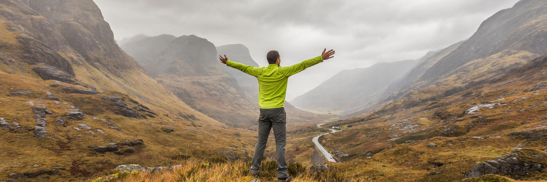 Rückansicht eines Mannes mit ausgestreckten Armen, der in einem Tal steht und auf die in Nebel gehüllten Berge blickt