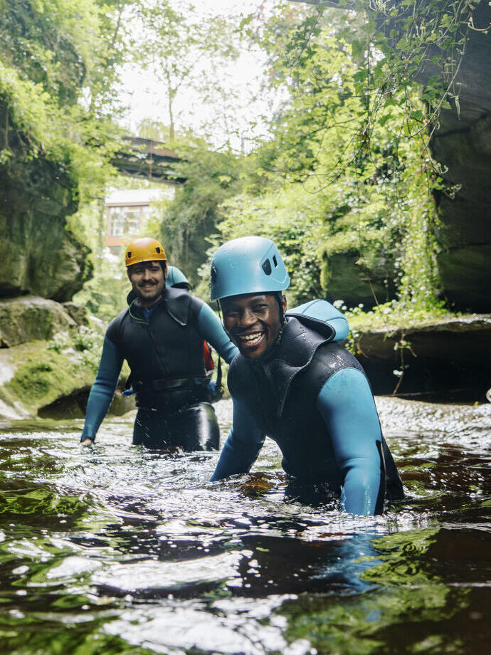 Two men wearing wetsuits and helmets wade waist deep in a gorge