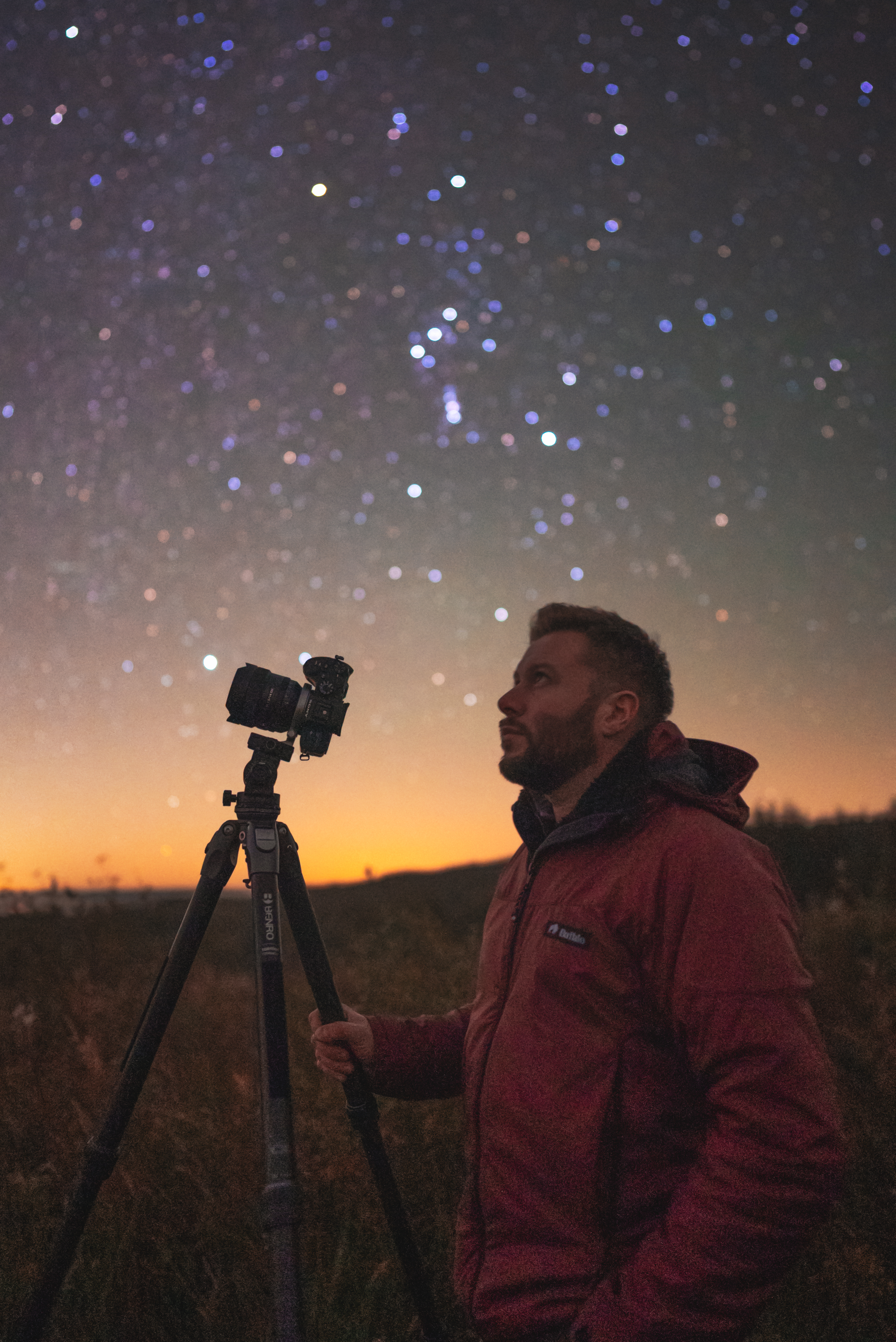 A person in a red jacket stands next to a camera on a tripod under a starry night sky, with a glowing orange horizon in the background.
