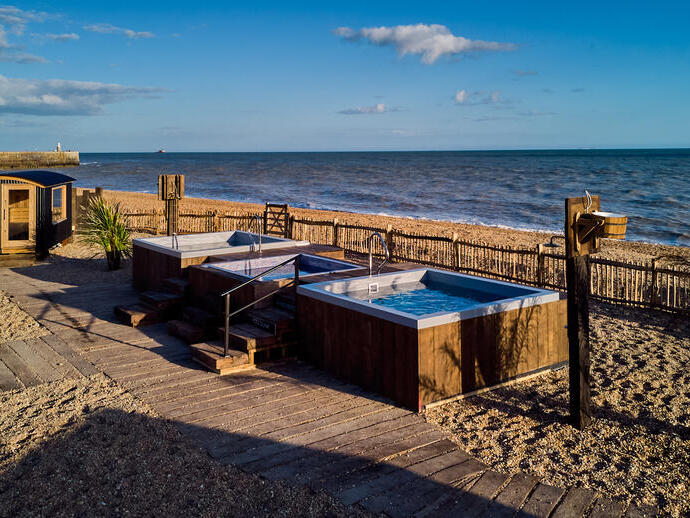Wooden hot tubs and a small sauna cabin on a sandy beach, overlooking the sea under a clear blue sky.