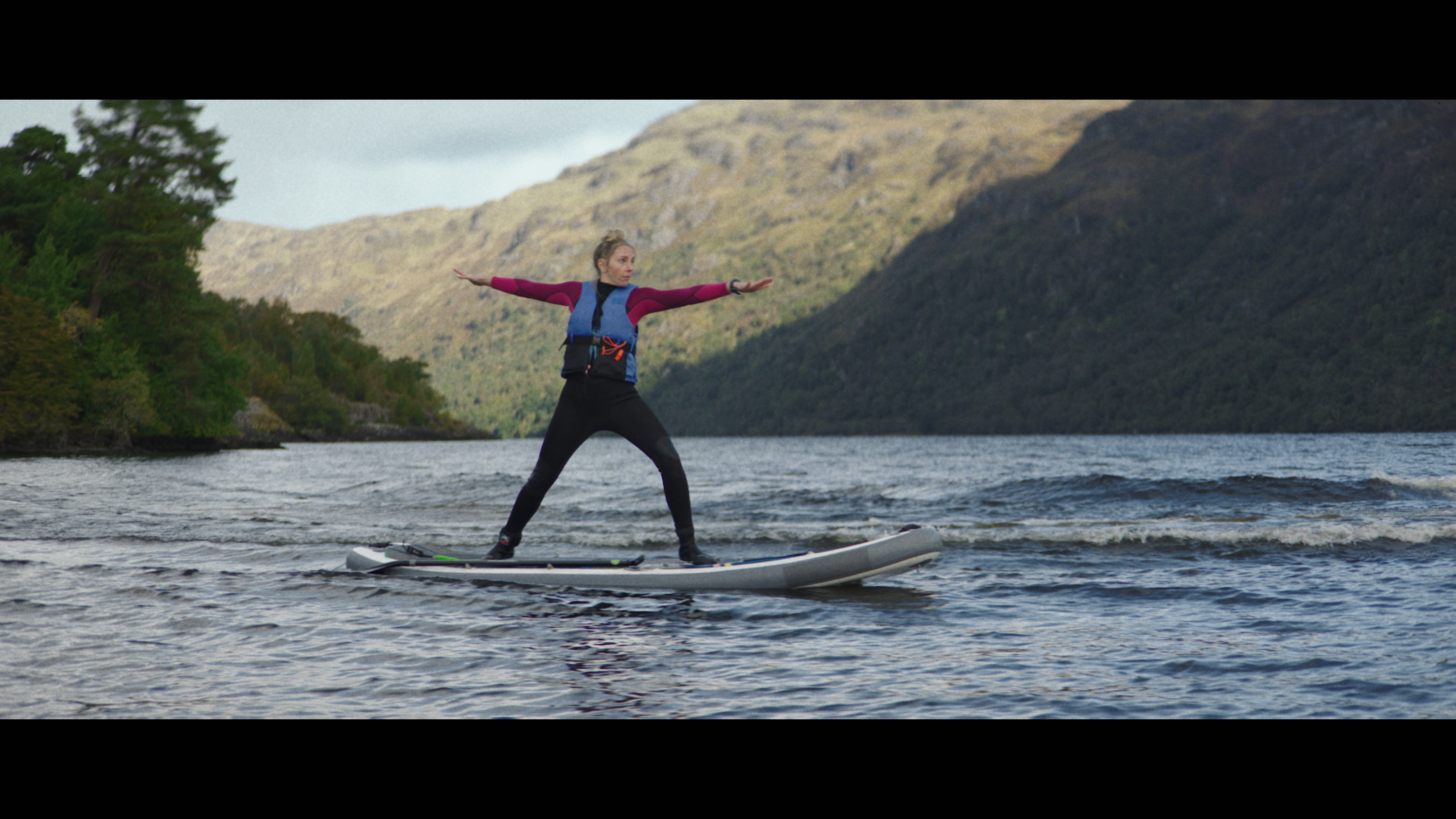 A woman stands on a paddleboard on a loch with mountains behind